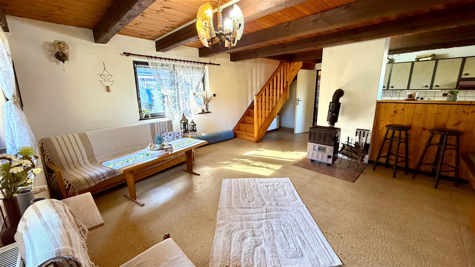 Světlý obývací pokoj s dřevěným stropem, krbovými kamny a parketovou podlahou, vybavený pohodlným posezením a rustikálním nábytkem.