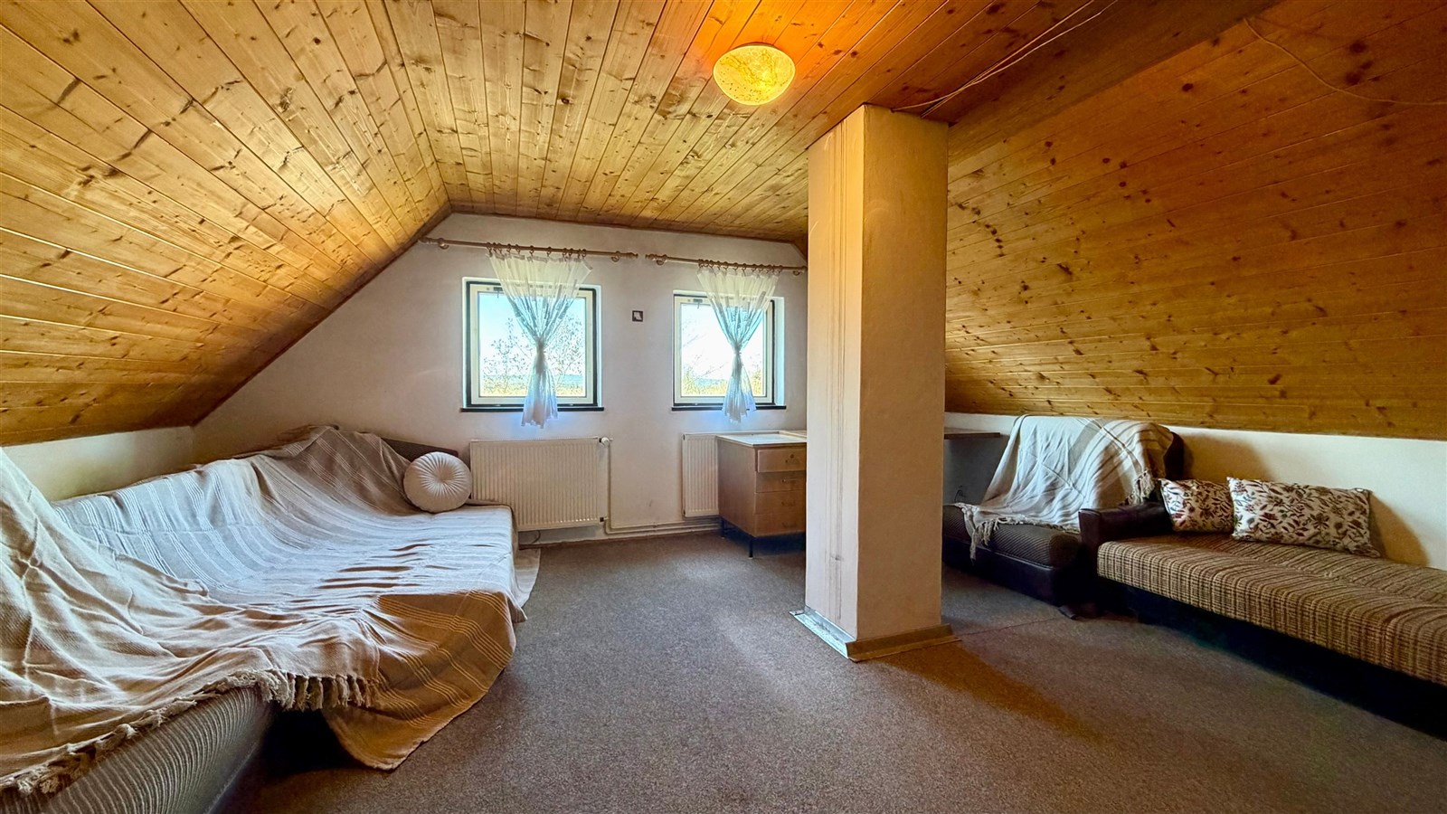 Podkrovní obývací pokoj s dřevěným stropem, velkými okny a světlým kobercem, útulný prostor vhodný k relaxaci.