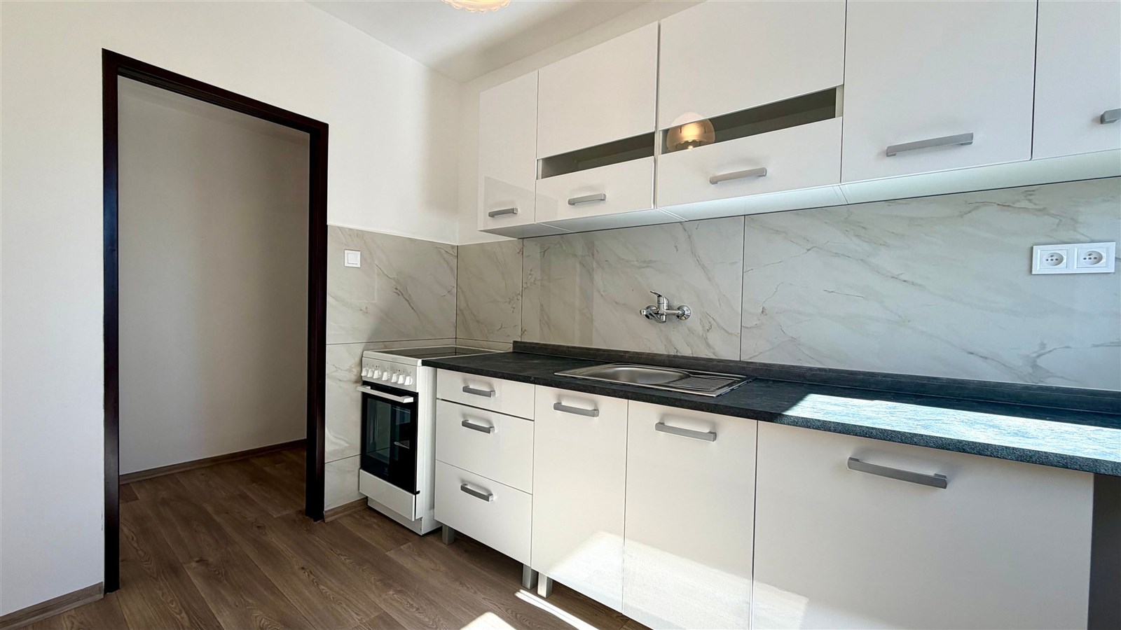 Moderní kuchyň s dřevěnou podlahou a mramorovým obkladem, vybavena bílými skříňkami a elektrickým sporákem, světlá a prostorná.