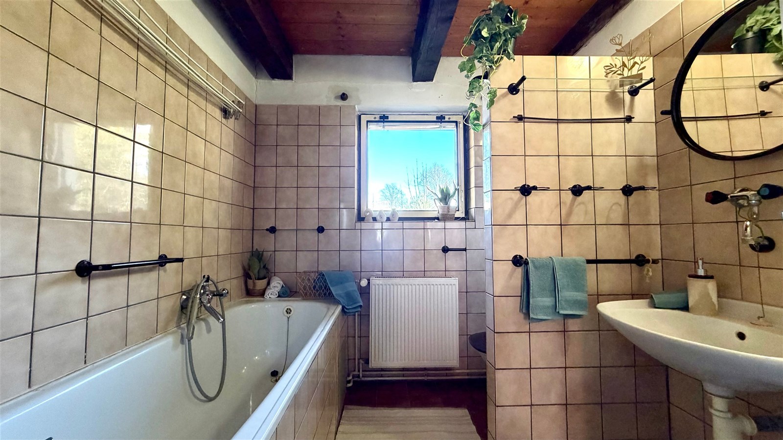 Koupelna s dlaždicemi, dřevěným stropem a velkým oknem, které zajišťuje dostatek světla. Obsahuje vanu a moderní vybavení.