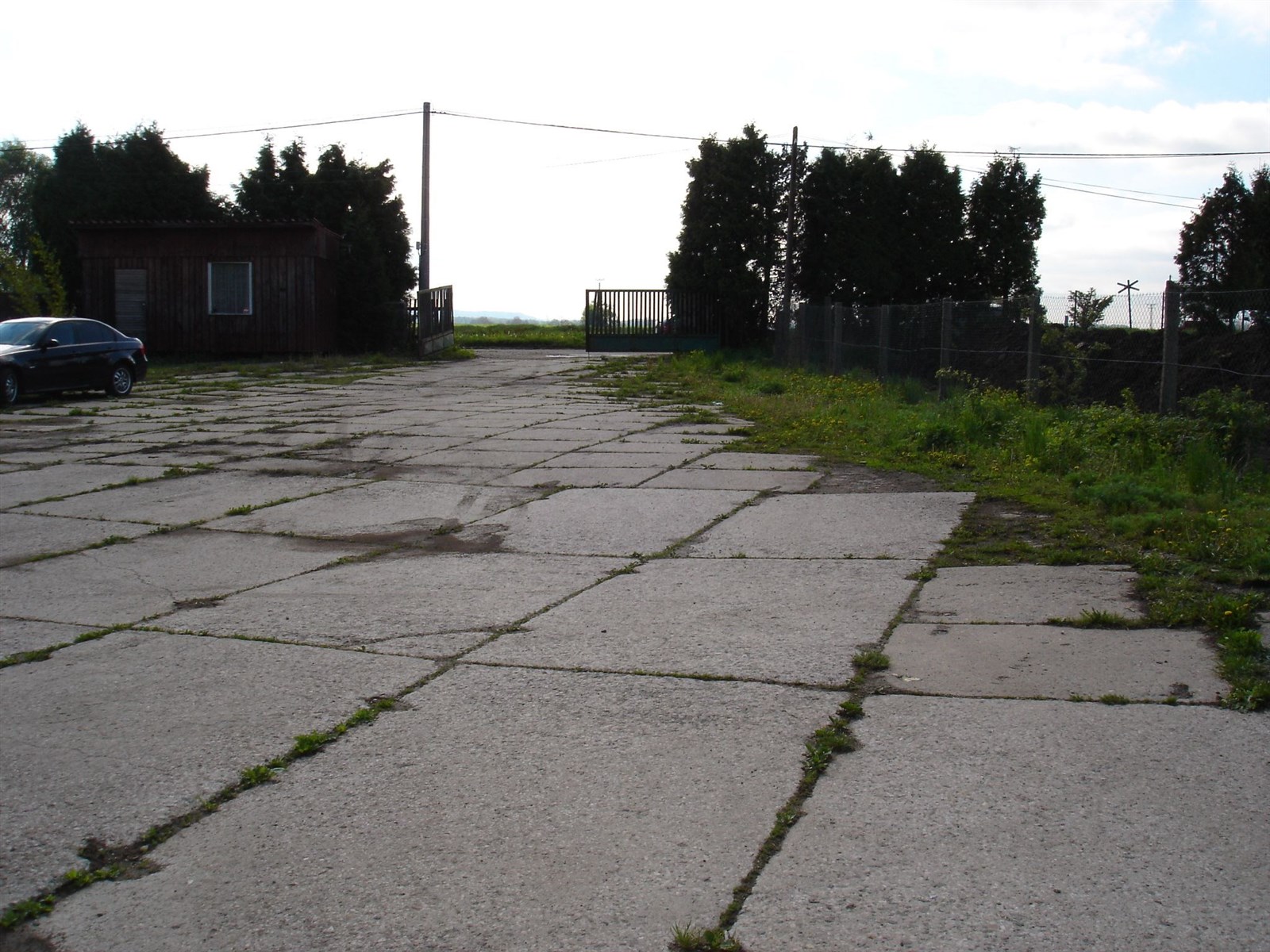 Zpevněná parkovací plocha s betonovým povrchem, oplocení z drátěného pletiva, mírně zarostlá vegetací, vhodné pro komerční využití.