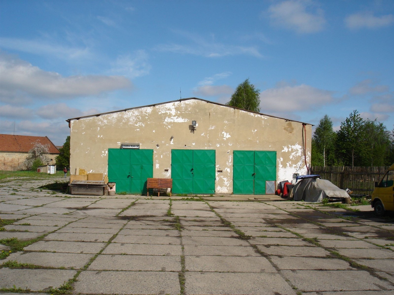 Průmyslová budova s betonovou plochou, zelenými vraty a opotřebenou fasádou, vhodná pro sklad nebo dílnu, na klidném místě.