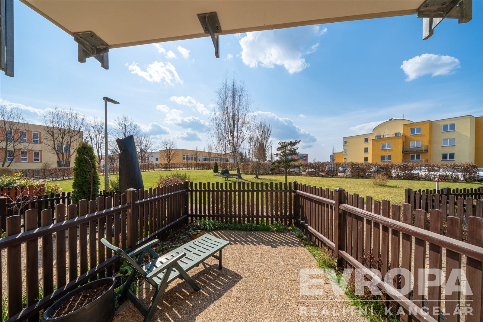 Terasa s dlažbou a výhledem do zahrady, obklopená dřevěným plotem, nabízí klidné prostředí v slunném bytovém komplexu.