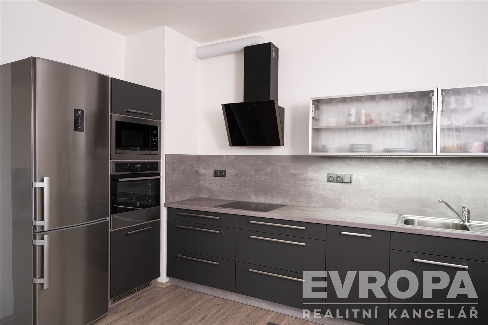 Moderní kuchyň s tmavou dřevěnou podlahou, vestavěné spotřebiče, stylová digestoř a šedé skříňky, minimalistický design.