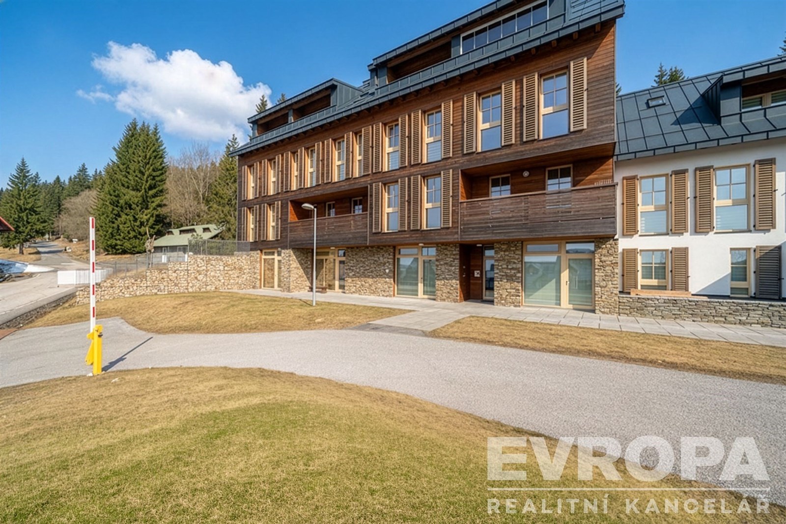 Moderní bytový dům s dřevěným obkladem, velkými okny a dobře udržovaným okolím, ideální pro milovníky klidného prostředí.