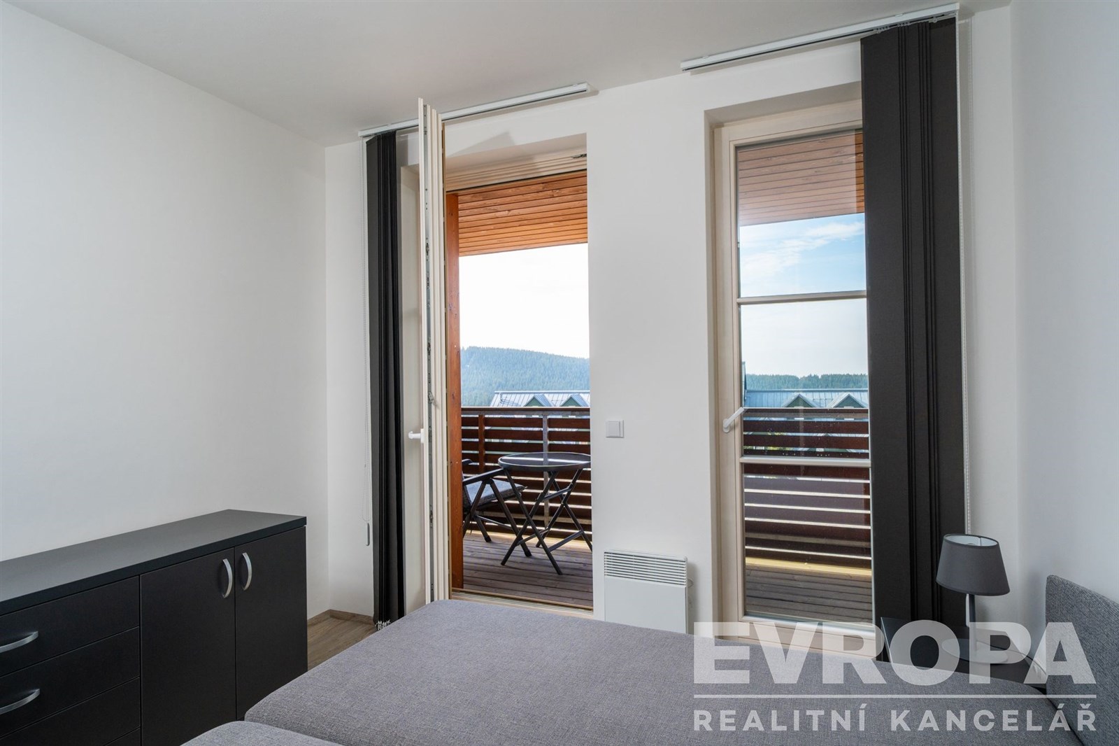 Prostorná ložnice s moderními velkými okny a parketovou podlahou nabízí vstup na balkon s malebným výhledem.