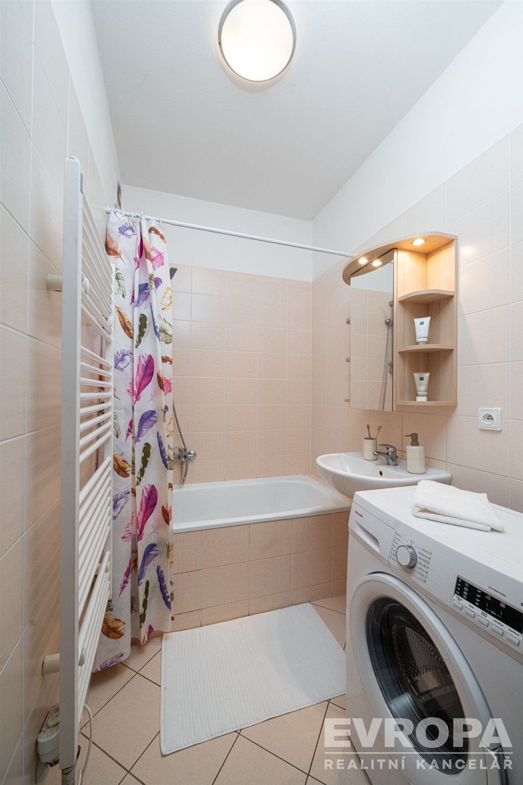 Koupelna s obklady v neutrálních tónech, vana, umyvadlo, moderní pračka a vzorovaný sprchový závěs.