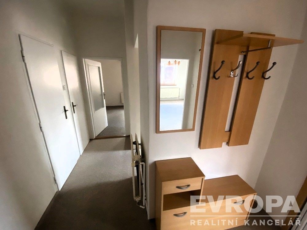 Vstupní hala se světlými stěnami a praktickým předsíňovým nábytkem. Moderní úložné prostory a zrcadlo, koberec pokrývá podlahu.