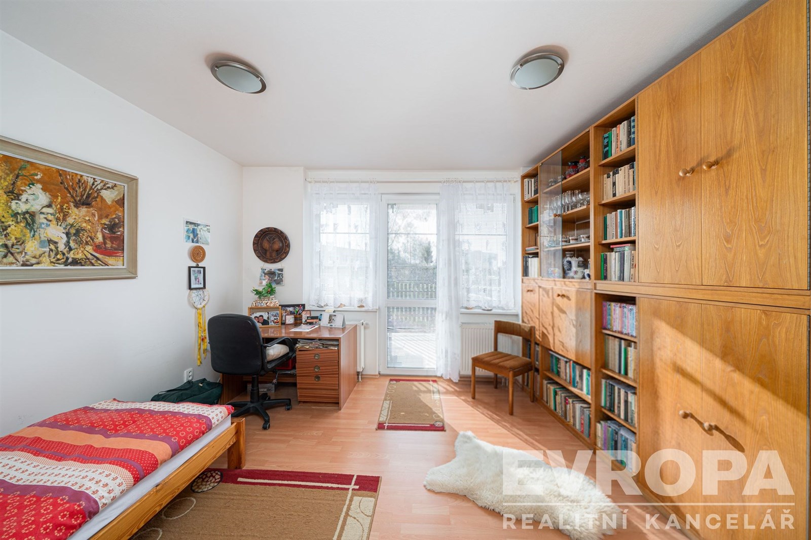 Světle zařízená pracovna s parketovou podlahou, vestavěnými skříněmi a velkými okny zajišťujícími dostatek denního světla.