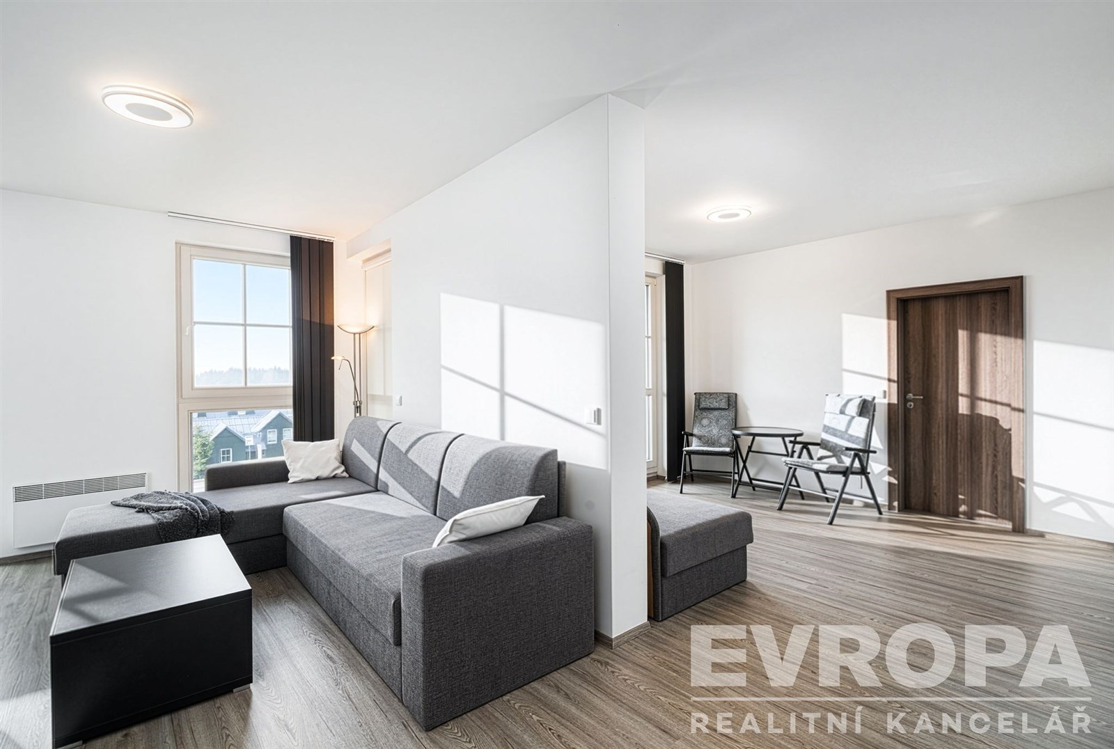 Světlý obývací pokoj s parketovou podlahou a moderním nábytkem, velká okna zajišťují přirozené světlo, stylový a minimalistický design.