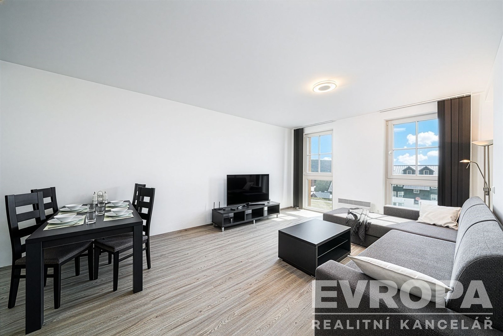 Světlý obývací pokoj s moderním nábytkem, parketovou podlahou a velkými okny, které zajišťují dostatek denního světla.