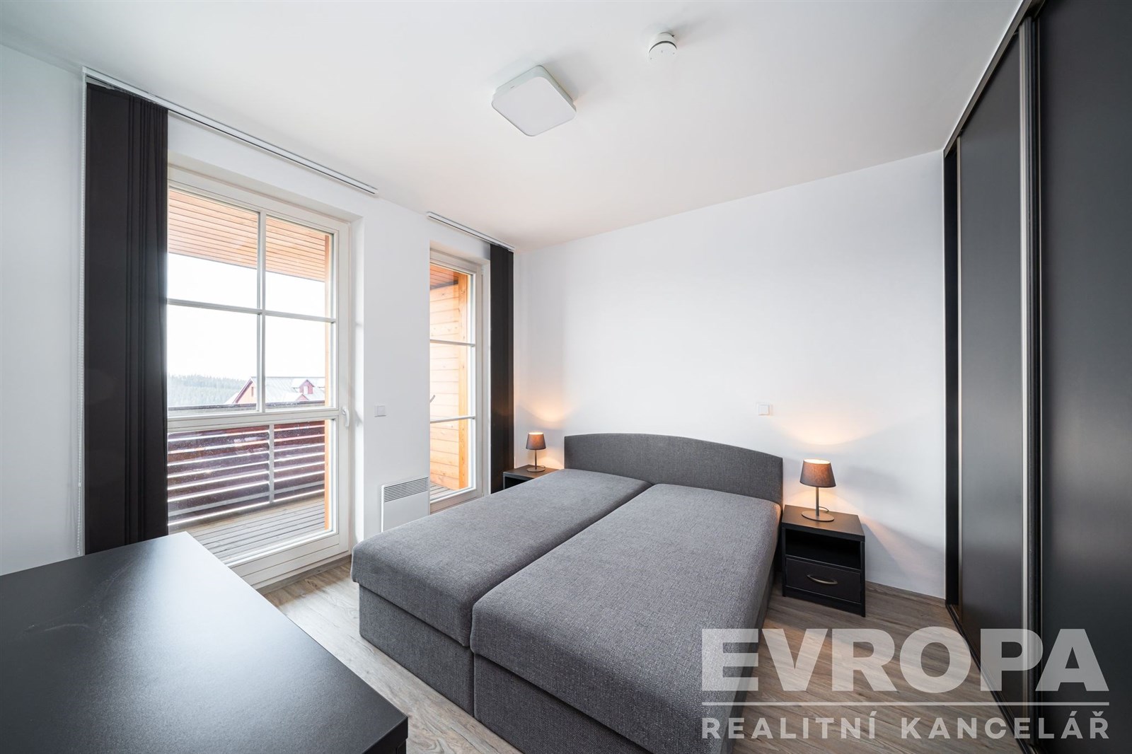 Prostorná ložnice s moderním nábytkem, velkými okny a vestavěnými skříněmi. Světlá výmalba a parketová podlaha dodávají elegantní vzhled.