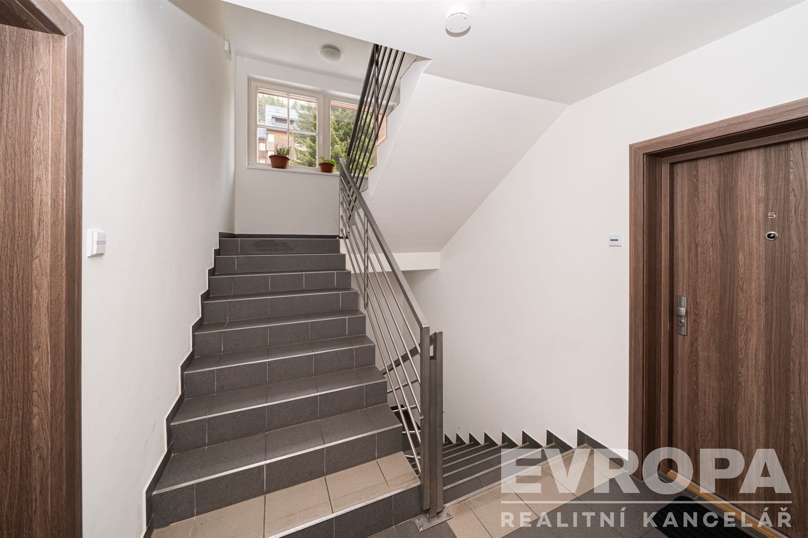 Schodiště s dlažbou, nerezovým zábradlím a moderními, světlými prvky, vedoucí k velkým oknům poskytujícím přirozené světlo