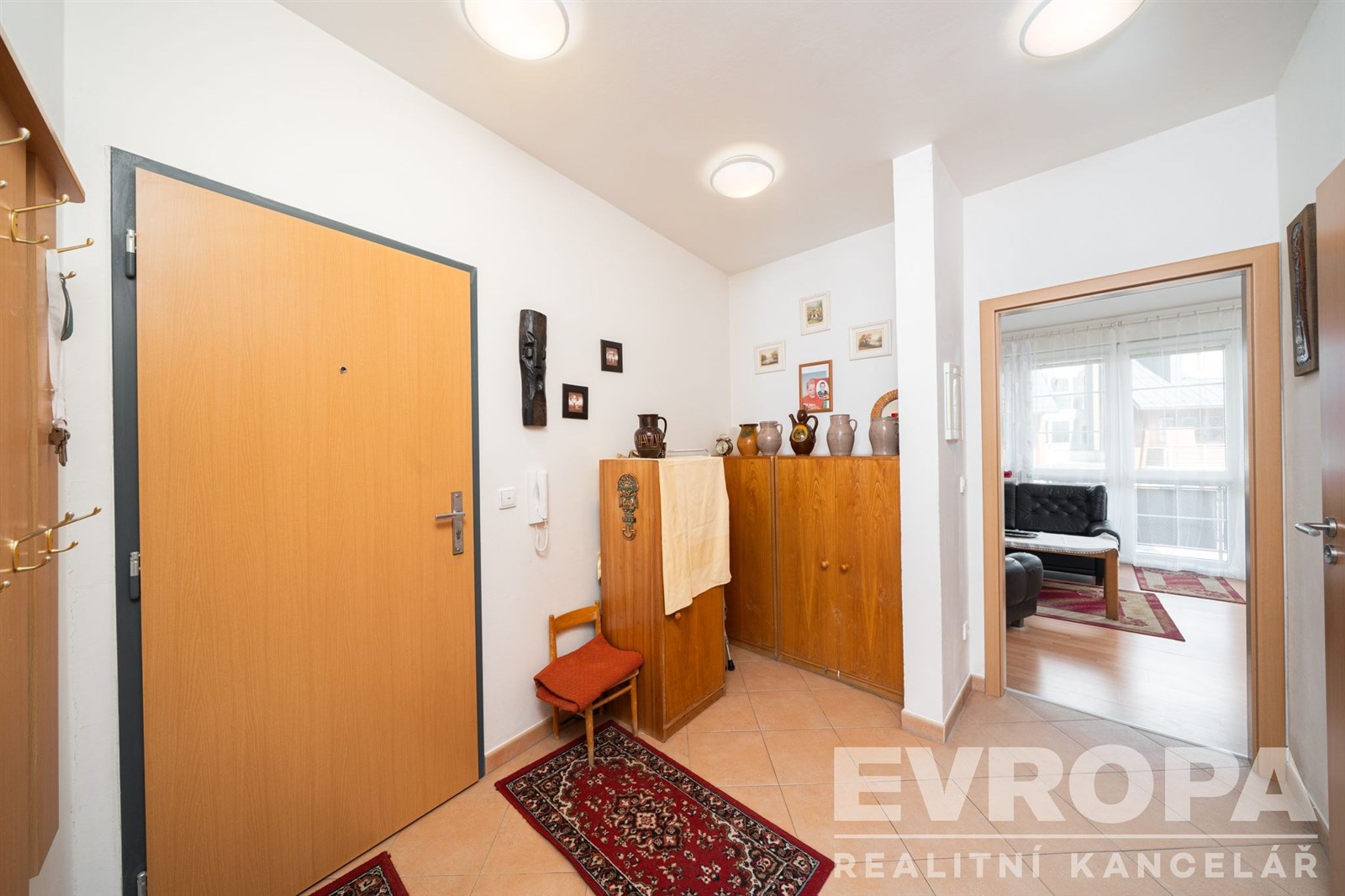 Vstupní hala s dlažbou, vestavěnými skříněmi a moderním osvětlením. Otevřený průchod do světlého obývacího pokoje se sedací soupravou.