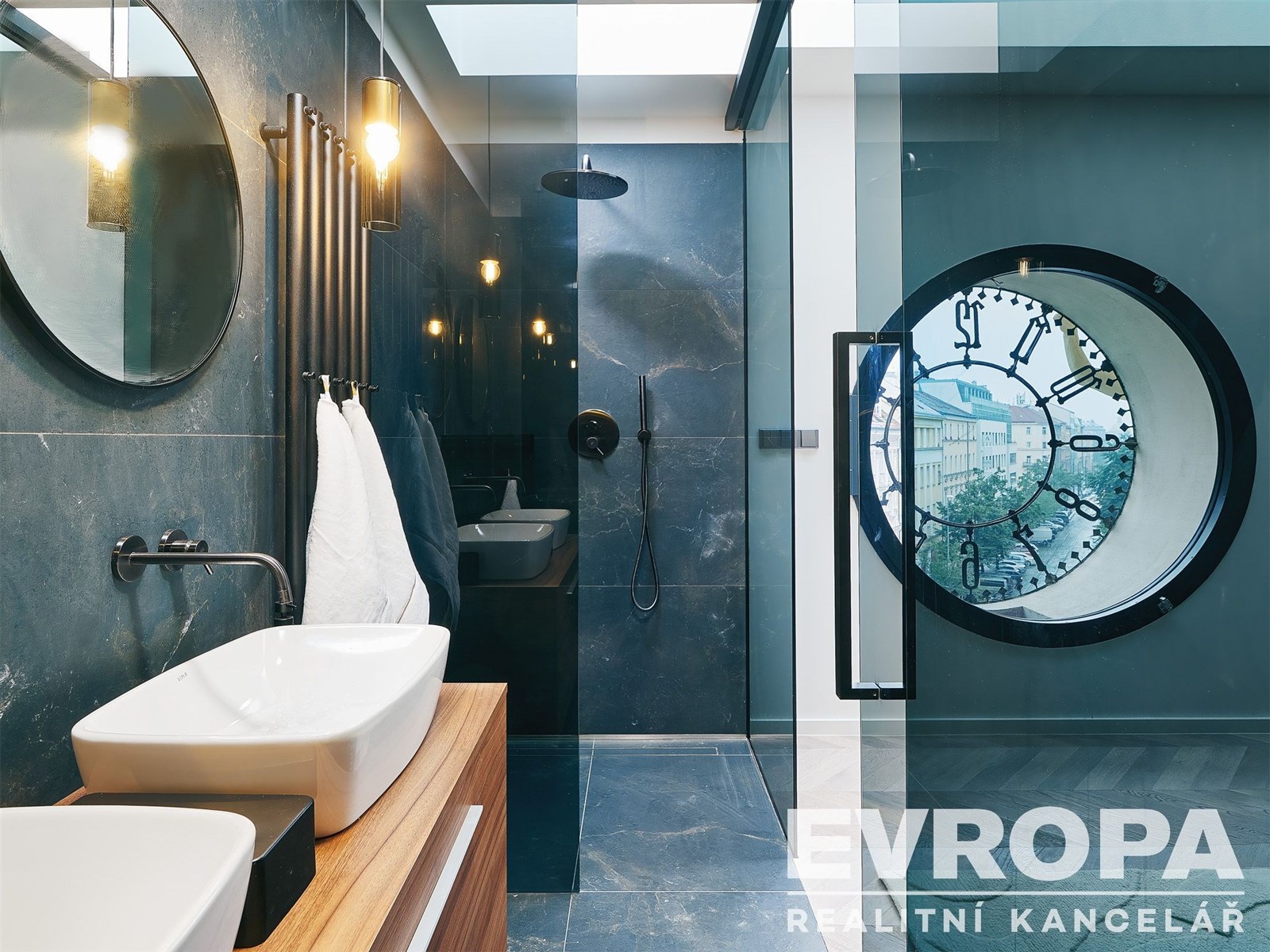 Moderní koupelna s tmavou dlažbou, velkým kulatým oknem a sprchovým koutem. Stylový design a elegantní umyvadlo na dřevěné desce.