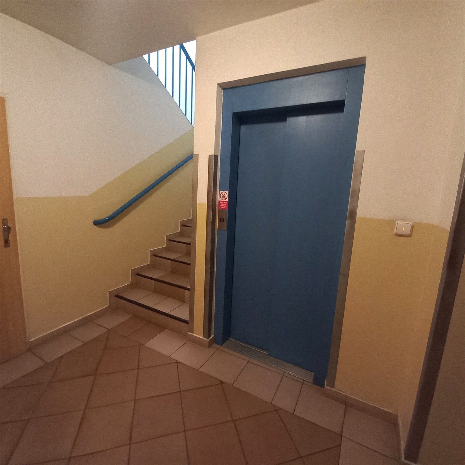 Vstupní hala bytového domu s dlažbou, modrým výtahem a schodištěm, čisté stěny a moderní design pro pohodlný přístup.