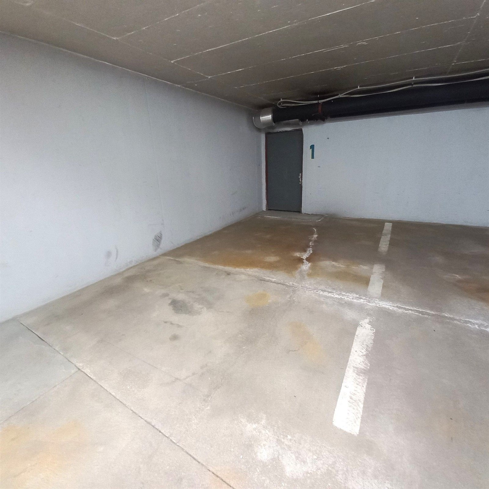 Prázdné parkovací stání v suterénu budovy se světlou omítnutou stěnou a betonovou podlahou, ideální pro bezpečné parkování