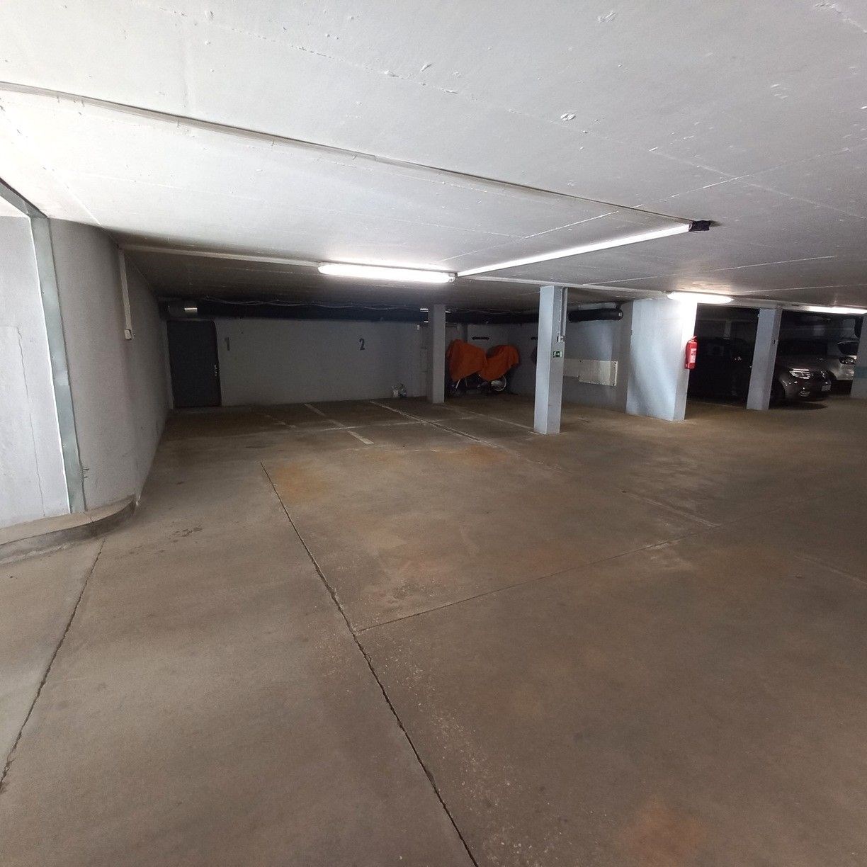 Prázdné parkovací místo v moderní garáži s betonovou podlahou, umělým osvětlením a světlými stěnami.