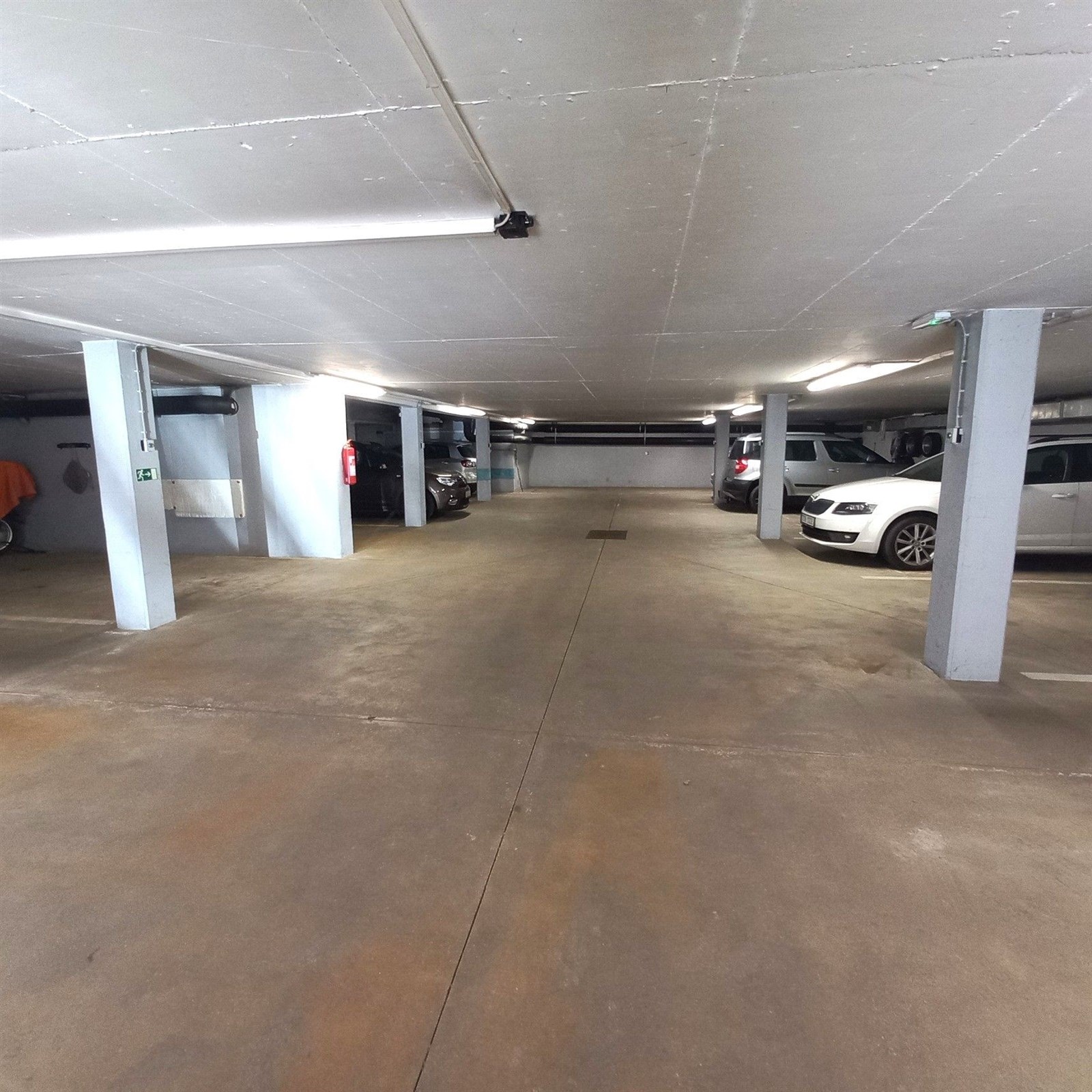 Podzemní garážové stání s betonovou podlahou, dostatečně osvětlené zářivkami, s několika zaparkovanými auty, nabízí bezpečné parkování.