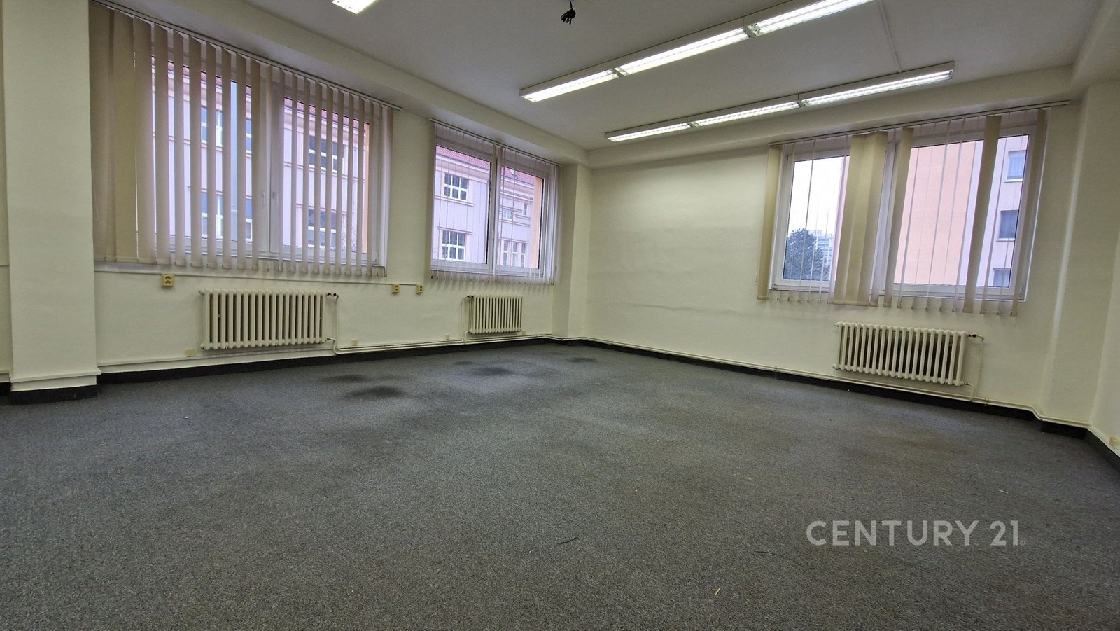 Prostorná kancelář s kobercovou podlahou, velkými okny a vertikálními žaluziemi, vhodná pro moderní pracovní prostředí.