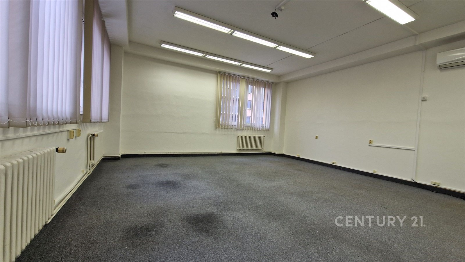 Kancelářský prostor s kobercovou podlahou, velkými okny a světlým stropem, ideální pro komerční využití, v dobrém stavu.