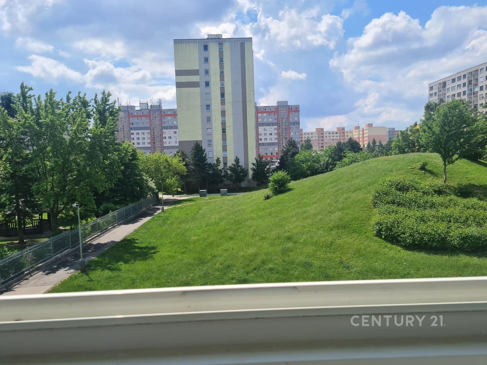 Výhled z okna na upravený zelený park v sídlišti s panelovými domy, modrá obloha, klidná lokalita, blízkost přírody a městské infrastruktury