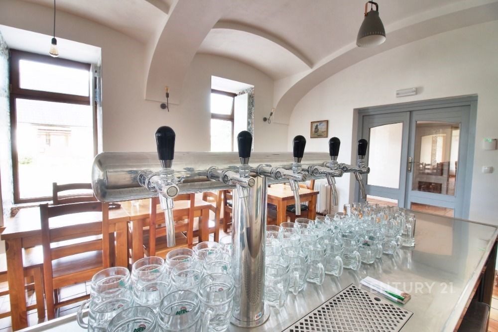 Prázdná místnost s dřevěnými stoly, velkými okny a výčepem, kde jsou umístěny pivní sklenice, prostor připravený pro akce nebo restauraci.