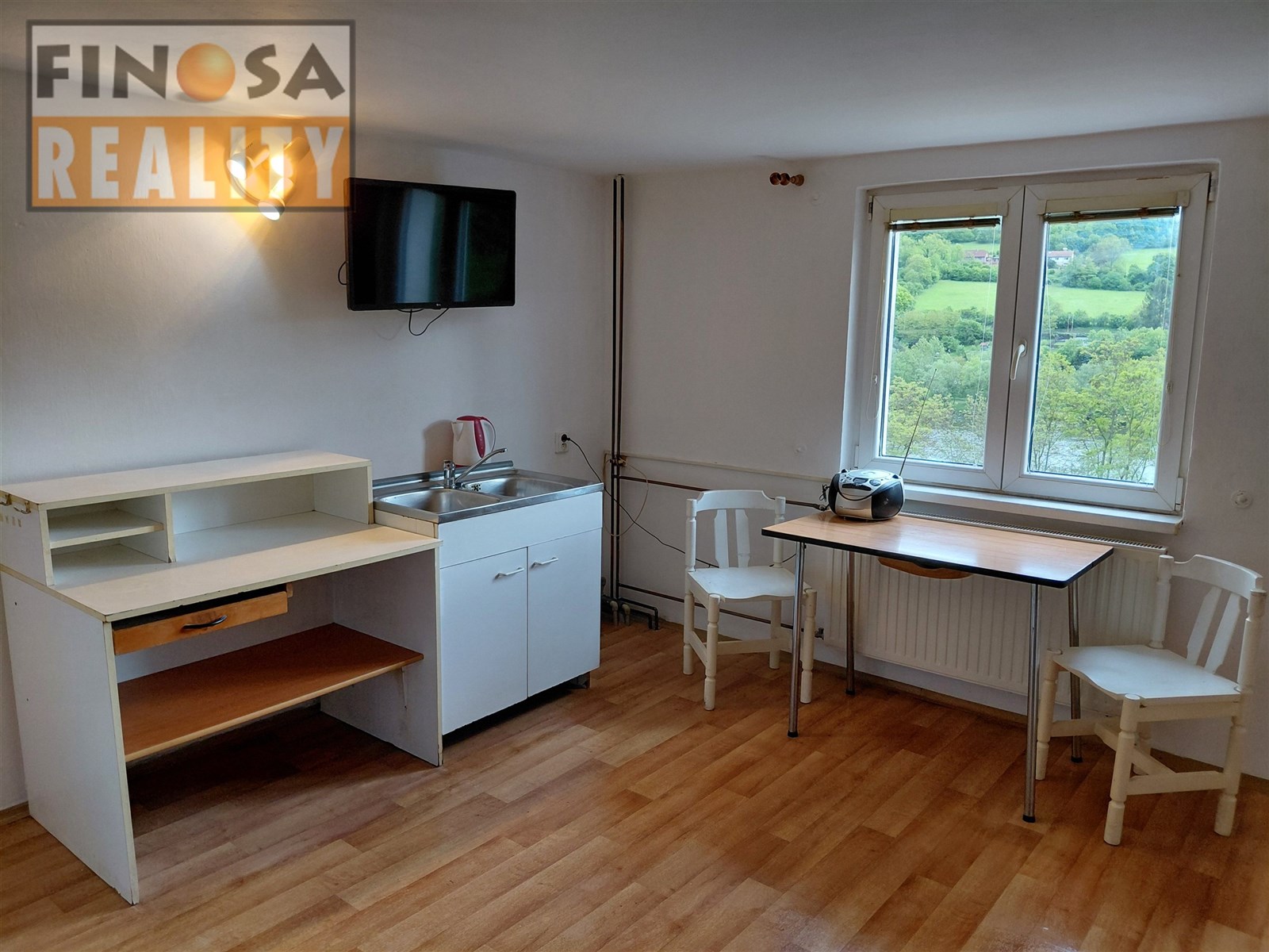 Malá kuchyňka s dřevěnou podlahou, velkým oknem a moderními doplňky, jako je televizor a osvětlení, nabízí útulný prostor k vaření.