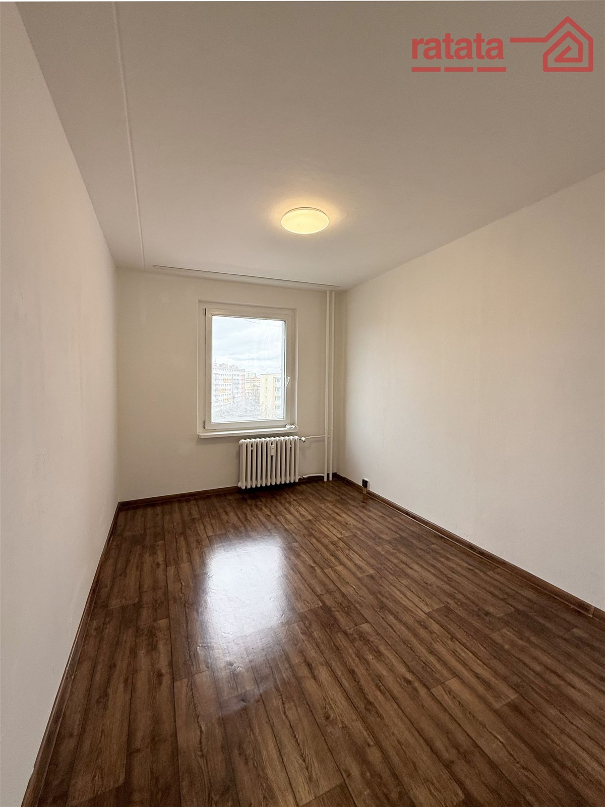 Světlá ložnice s parketovou podlahou, velké okno nabízí výhled na město, moderní úprava a minimalistický styl vytváří elegantní atmosféru.