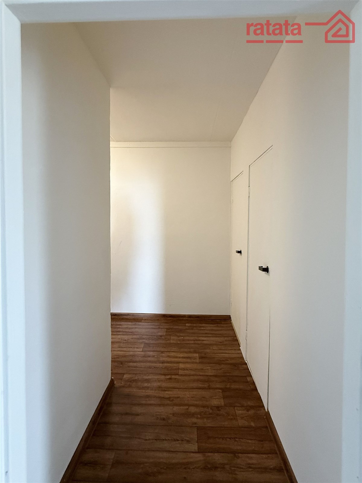 Úzká chodba s dřevěnou podlahou, bílými stěnami a moderními dveřmi, ideální pro vestavěné skříně nebo úložné prostory.