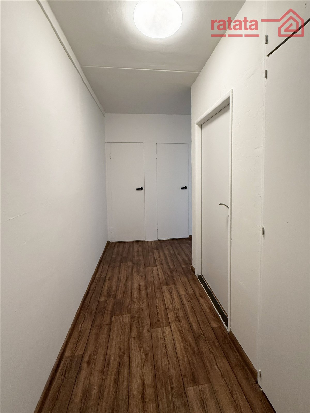 Úzká chodba s dřevěnou podlahou a bílými stěnami, moderní design, jednoduché čisté linie, dveře vedou do dalších místností.