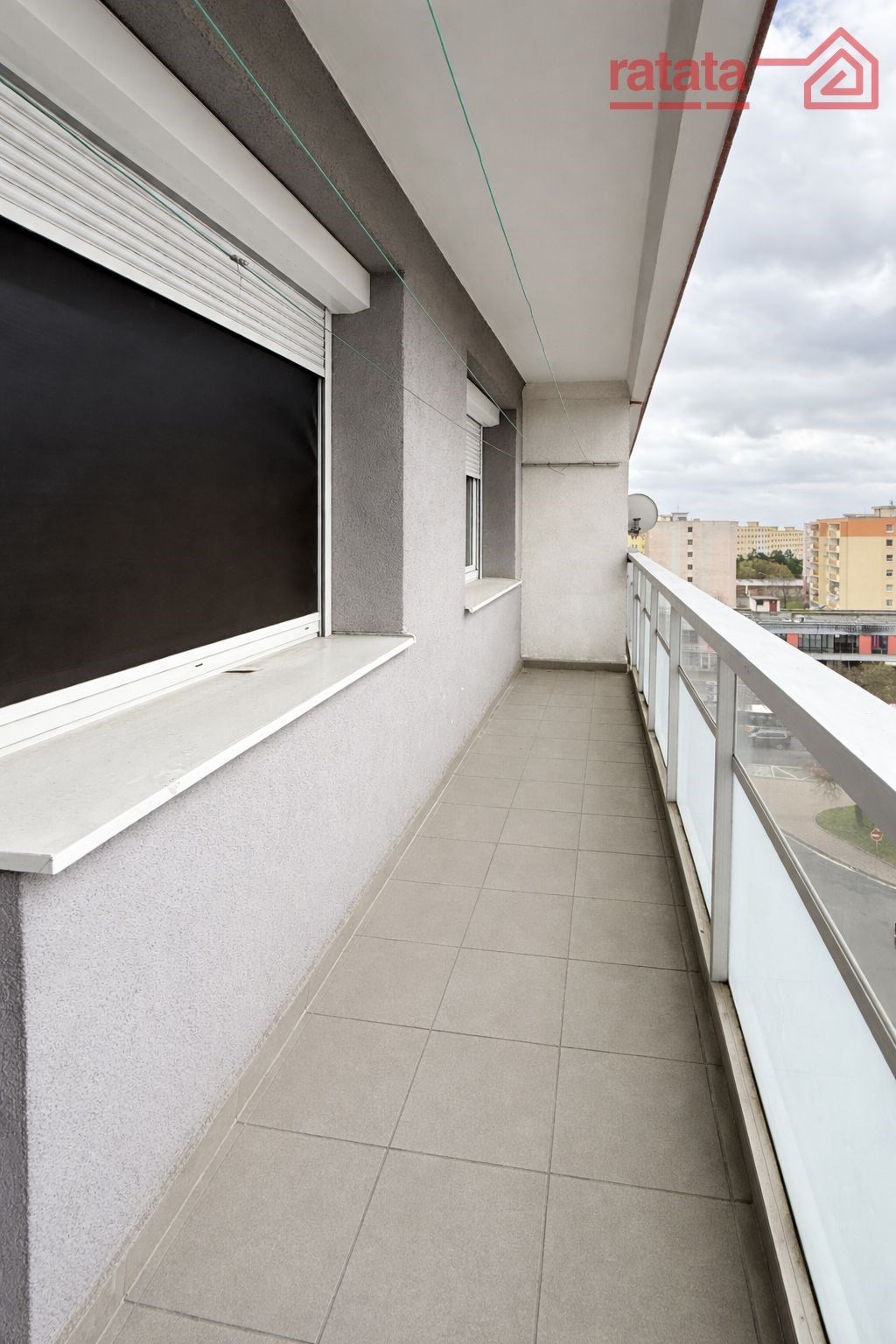 Dlouhý balkon s dlažbou, moderní zábradlí, výhled na město, udržovaný stav, ideální k relaxaci.