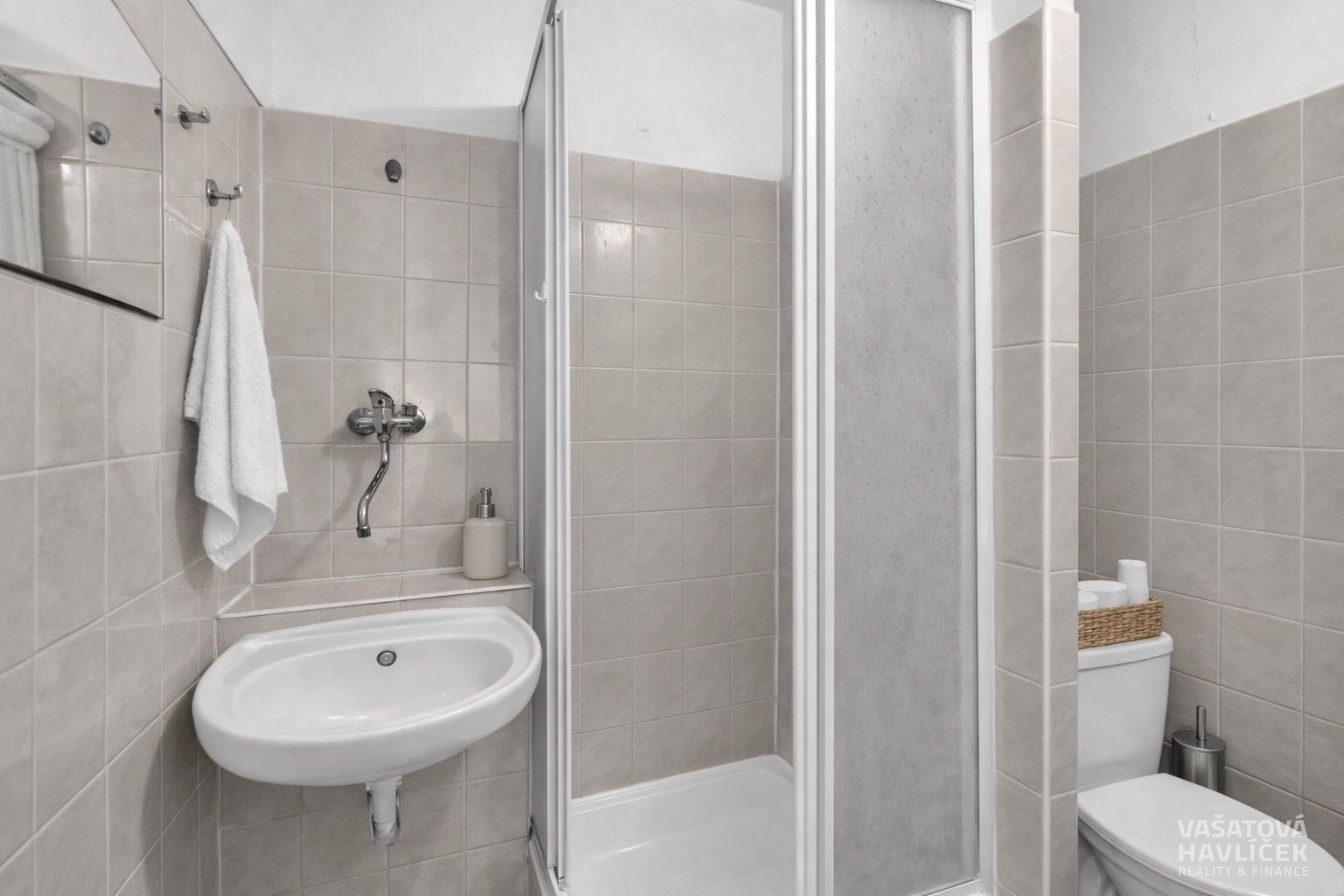 Moderní koupelna s dlažbou, sprchovým koutem a bílým umyvadlem, světlé stěny dodávají prostoru vzdušnost a čistotu.