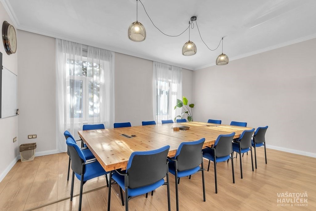 Zasedací místnost s dřevěným stolem a parketovou podlahou, velká okna zajišťují dostatek světla, moderní design osvětlení.