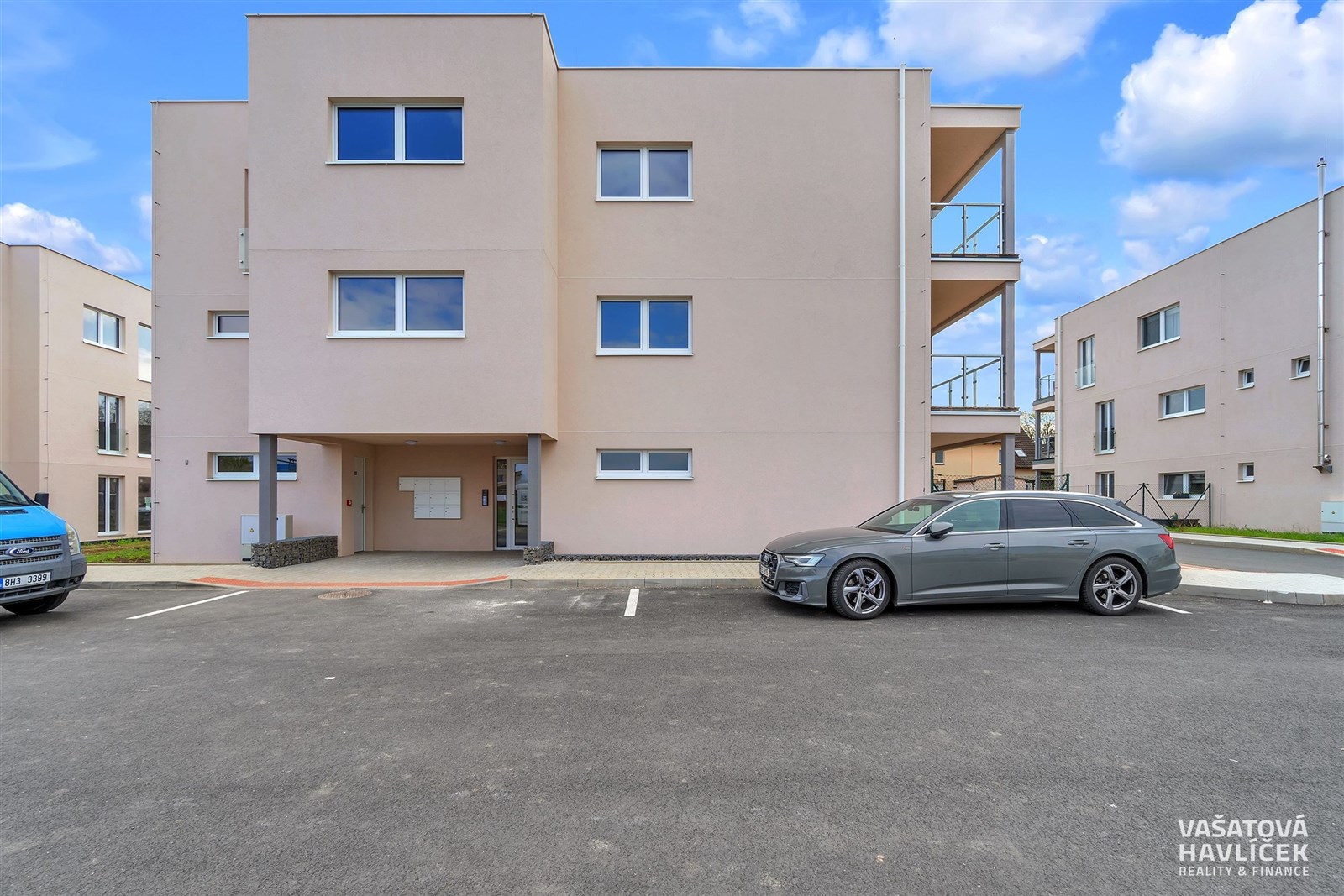 Třípodlažní bytový dům s moderní fasádou, parkoviště s asfaltovým povrchem, blízkost městské infrastruktury, novostavba v klidné lokalitě