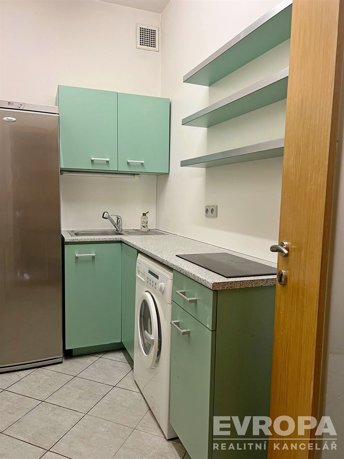 Kompaktní kuchyň s dlažbou a pastelově zelenými skříňkami, vybavená moderním spotřebiči a vestavěnými policemi pro úložný prostor.