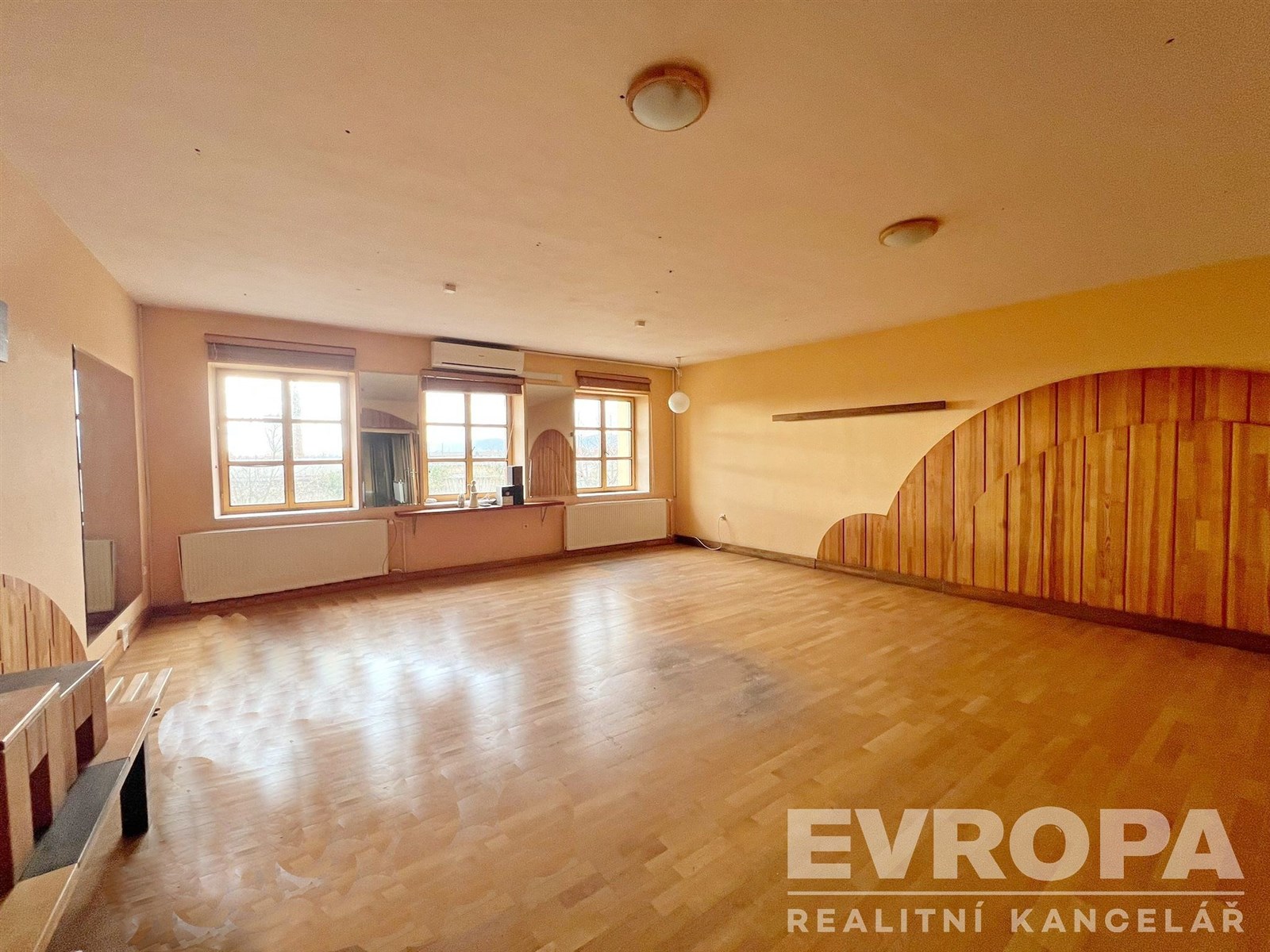 Prostorný obývací pokoj s parketovou podlahou, velká okna umožňují dostatek přirozeného světla, dřevěné obložení stěn vytváří útulnou atmosféru