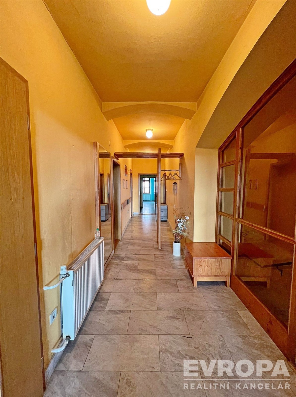 Dlouhá chodba s dlažbou, dřevěnými dveřmi a vestavěnými skříněmi, v rekonstruovaném stylu, nabízí přístup do několika místností.