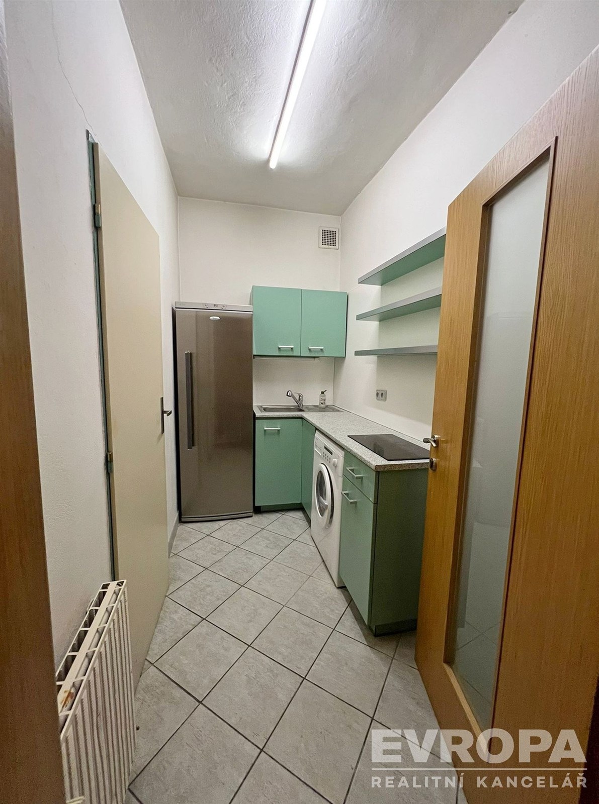 Úzká kuchyň s dlažbou, zelenými skříňkami a moderními spotřebiči, zahrnující pračku a nerezovou lednici, vhodná pro praktické využití.