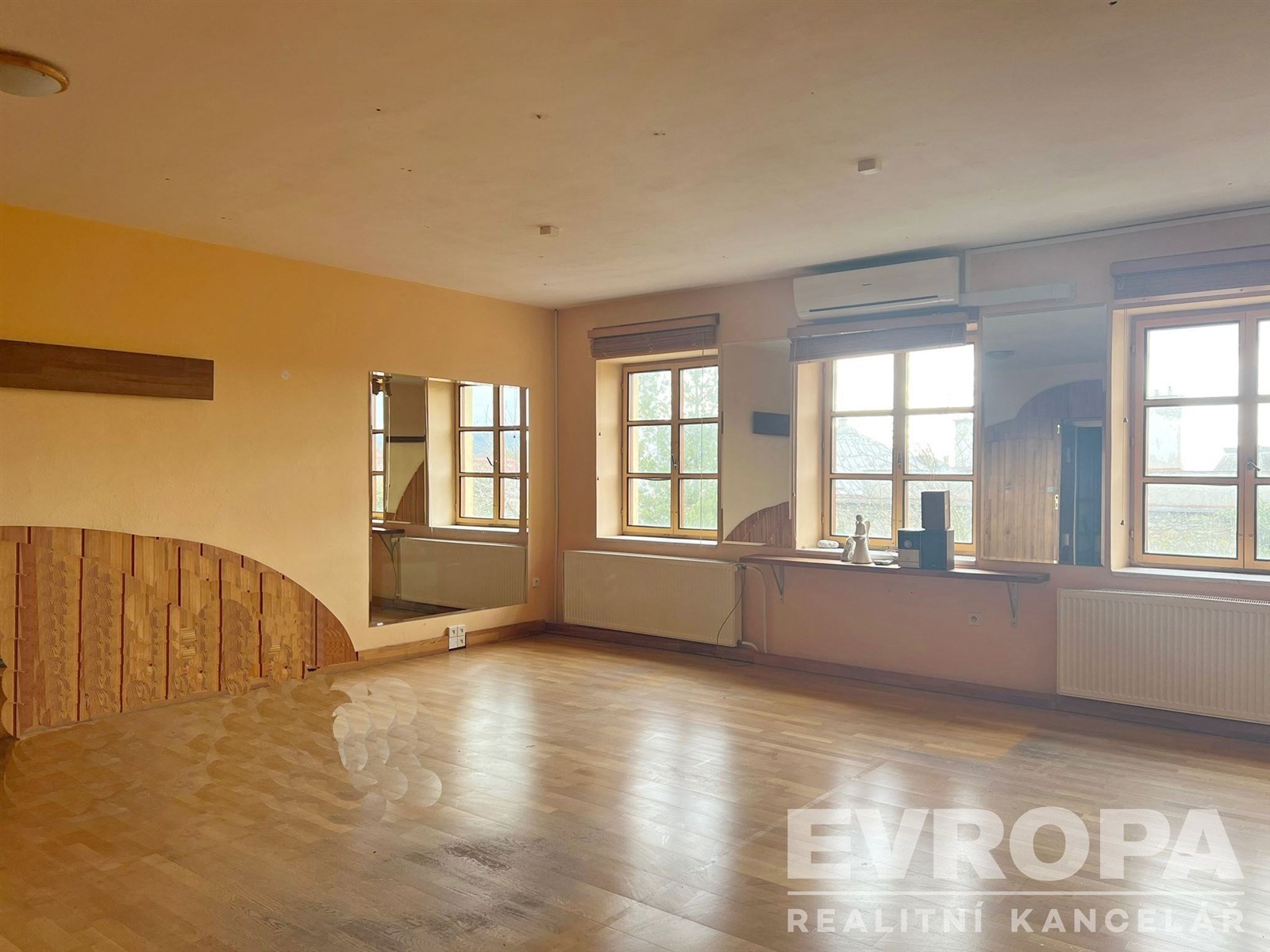 Prostorný obývací pokoj s dřevěnou parketovou podlahou, velkými okny a světlým designem, vytváří útulnou atmosféru.