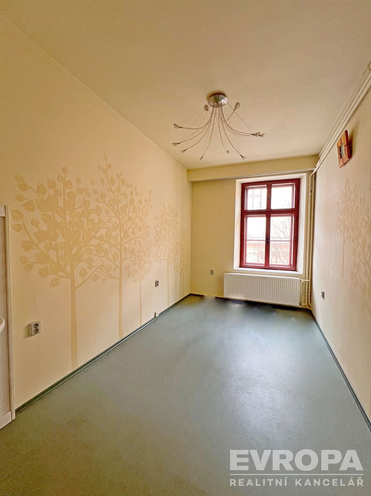 Prázdná místnost s malbou stromů na světle žluté stěně, velké červeně orámované okno zajišťuje přirozené světlo, světlý prostor k úpravě.