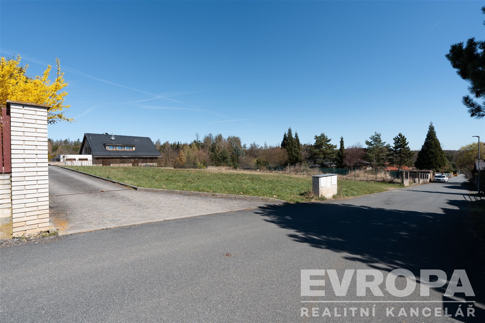 Prázdný pozemek s asfaltovou cestou, blízkostí zeleně a vzrostlými stromy, nacházející se v klidné oblasti vhodné pro výstavbu.