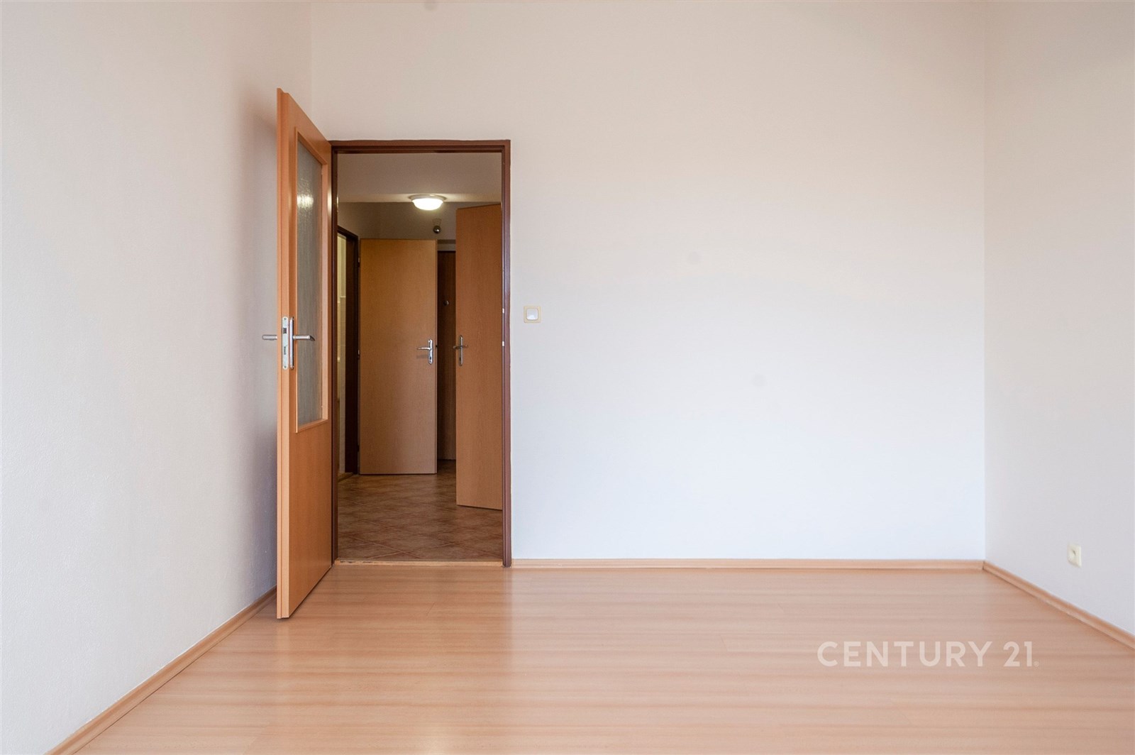 Prázdný pokoj s dřevěnou podlahou, bílými stěnami a dveřmi vedoucími do předsíně, ideální pro moderní zařizování.