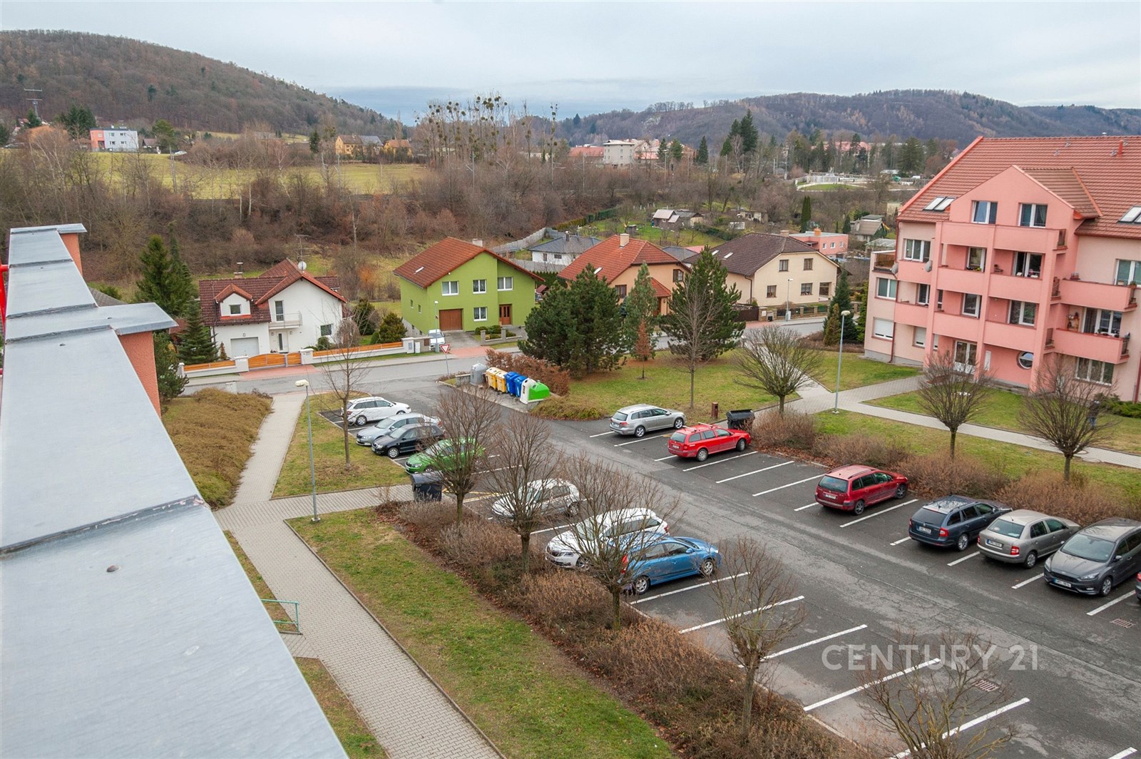 Výhled z balkonu na klidnou rezidenční čtvrť s rodinnými domy a parkovacími místy, okolní zeleň a kopcovitá krajina v pozadí, prostorné parkoviště.