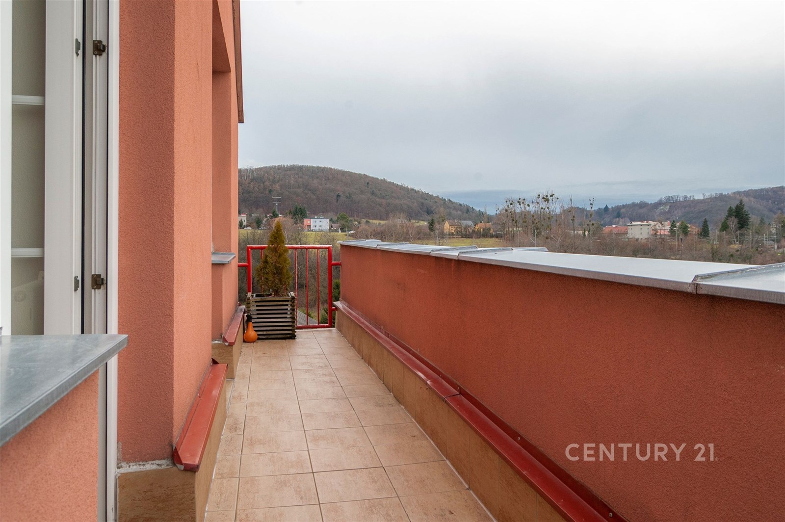 Balkón s dlažbou, červené zábradlí lemované rostlinami, nabízí panoramatický výhled na okolní krajinu a vytváří ideální prostor pro relaxaci.