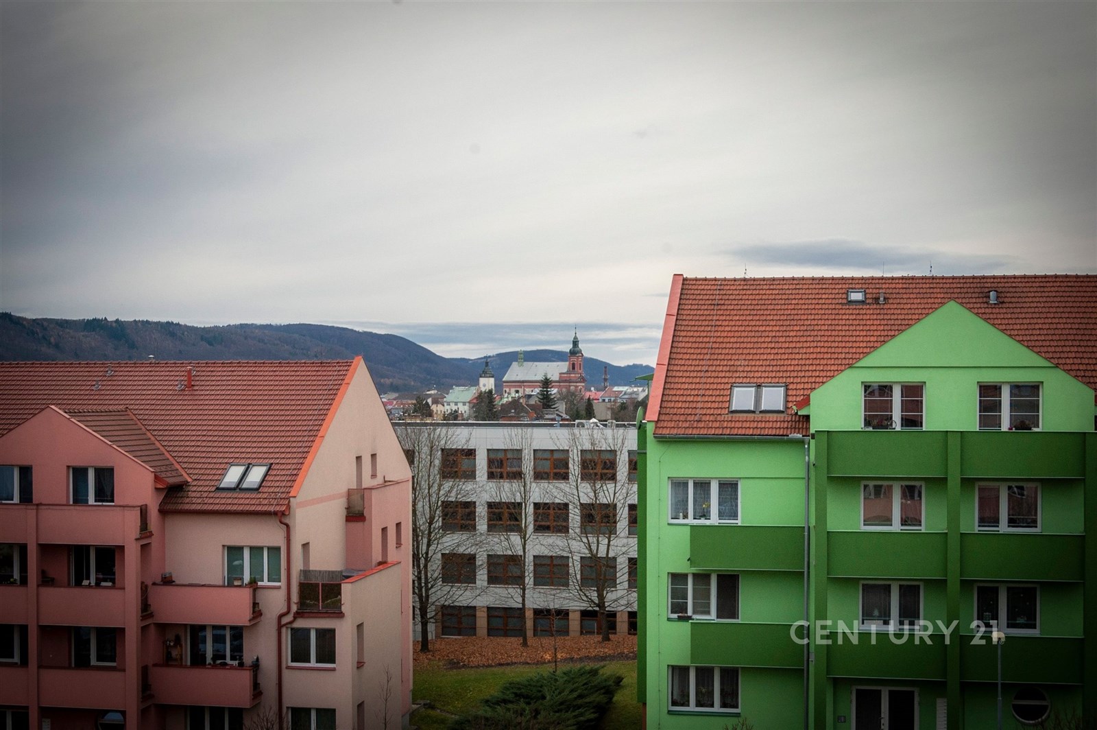 Barevné panelové domy s červenými střechami a výhledem na městské panorama na pozadí, připomínající rekreační lokalitu s klidným prostředím.