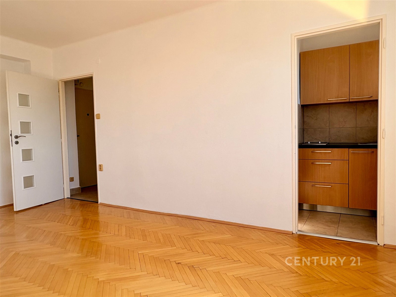 Světlý obývací pokoj s parketovou podlahou a moderními dřevěnými dveřmi, vedle kuchyň s vestavěnými skříněmi a dlažbou, ideální pro renovaci.