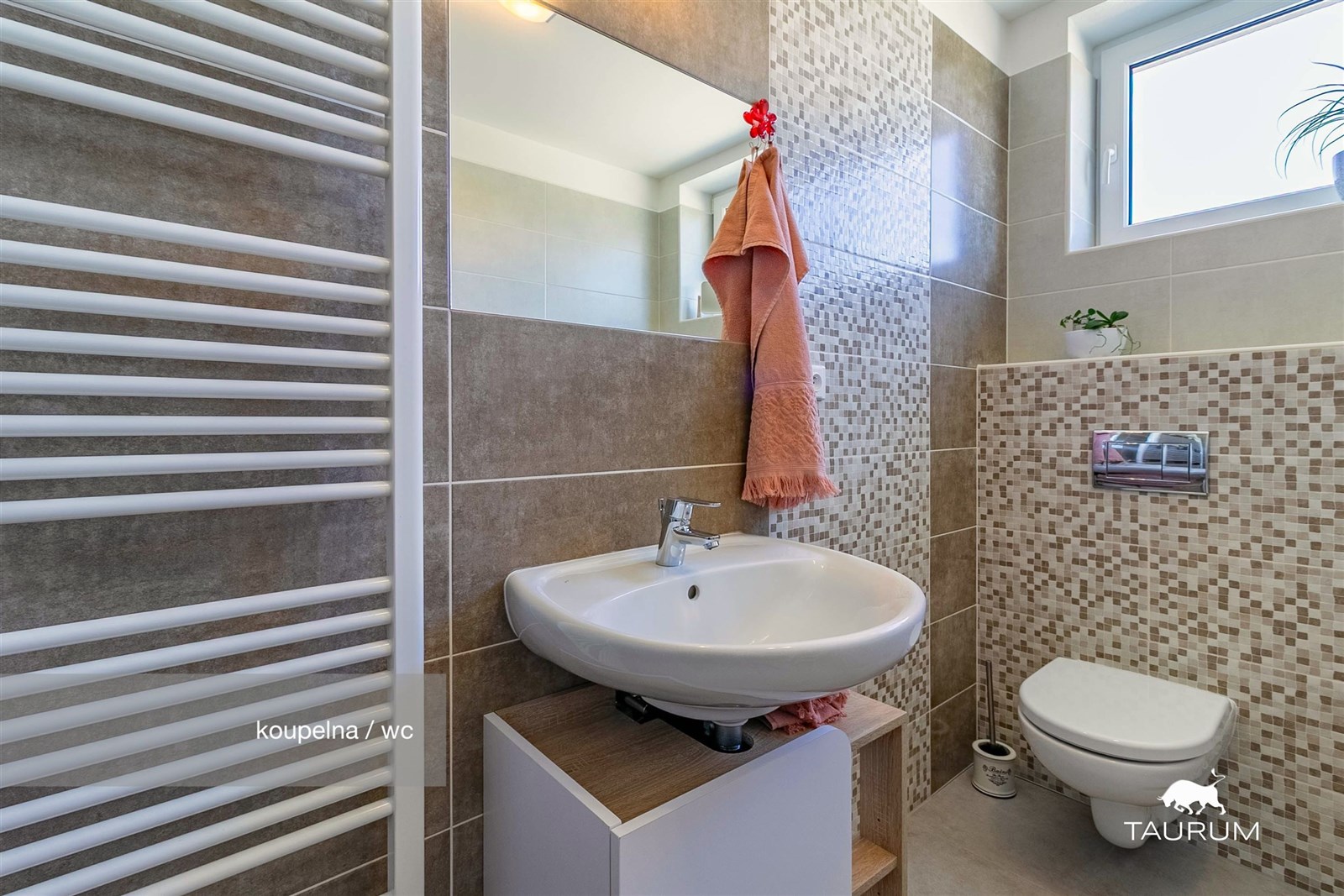 Moderní koupelna s keramickými dlaždicemi, závěsnou toaletou a umyvadlem, světlá díky přítomnosti okna.