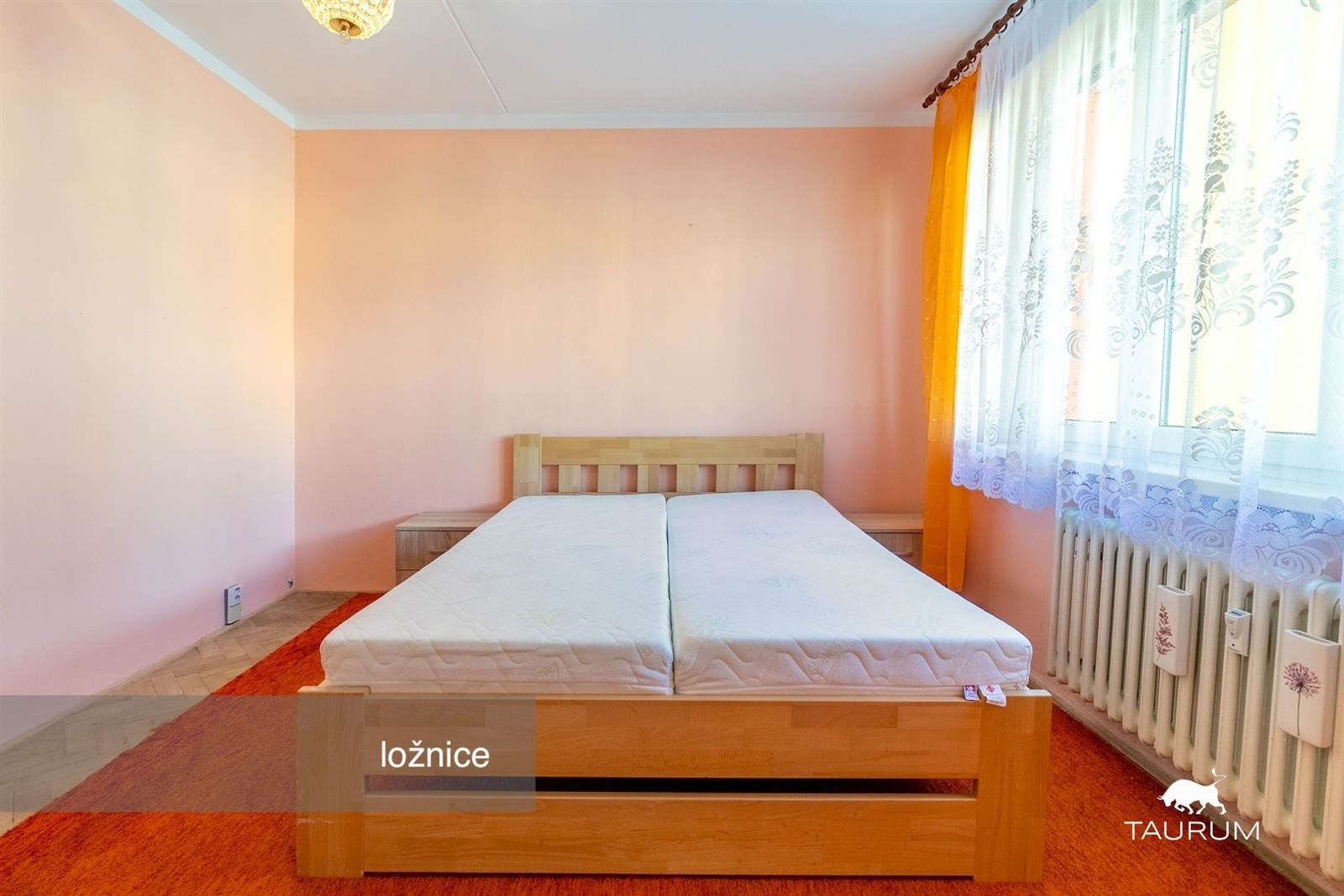 Prostorná ložnice s dřevěnou postelí, velká okna s krajkovými záclonami, světle růžové stěny, útulný vzhled, ideální pro rodinu.