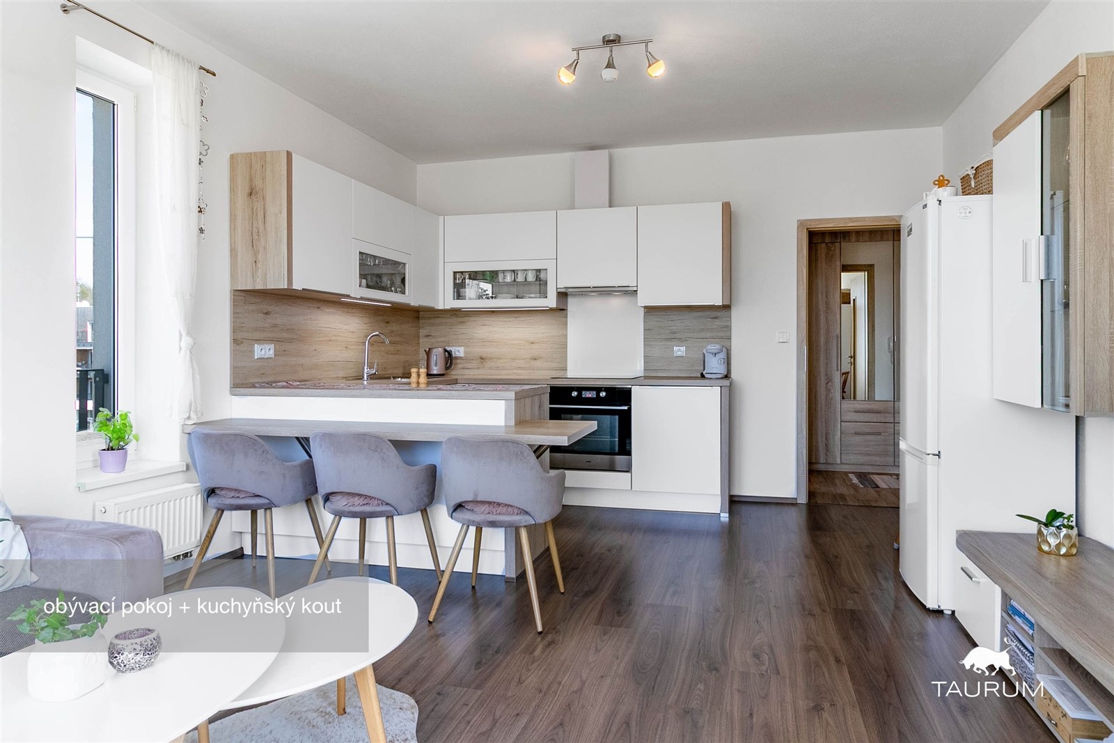 Moderní kuchyň s parketovou podlahou, bílými skříňkami a velkým oknem propojená s obývacím prostorem, nabízí světlé a vzdušné prostředí.